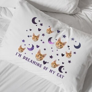 May include: White pillowcase with a design of orange tabby cat faces, purple crescent moons, stars, and hearts. The phrase "I'M DREAMING OF MY CAT" is printed in purple. The pillowcase is on a white bed.