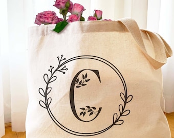 May include: A white canvas tote bag with a black floral design and the letter "C" in the centre. The bag has two handles.