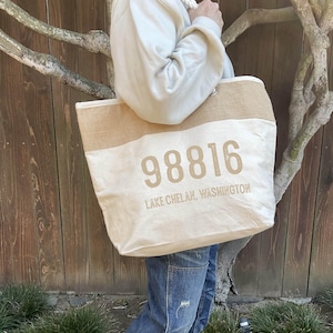 May include: A large, white canvas tote bag with brown jute trim. The bag has a brown, distressed print of the number "98816" and the text "LAKE CHELAN, WASHINGTON".