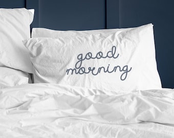 May include: White bed linens with a white pillow with the text "good morning" embroidered in blue.