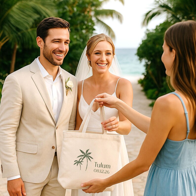May include: A beige tote bag with a palm tree graphic and the word "Tulum" and the date "08.12.26" printed on it. The bag is being held by a person in a blue dress, and is being offered to a smiling couple, a bride and groom, on a path.