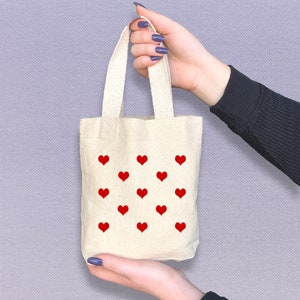 May include: A small, white canvas tote bag with red heart designs. The bag has two handles and is being held by a hand.