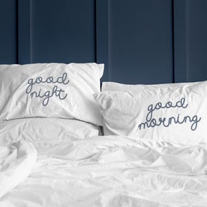 May include: Two white pillowcases with blue embroidered text. The pillowcase on the left says "good night" and the pillowcase on the right says "good morning".