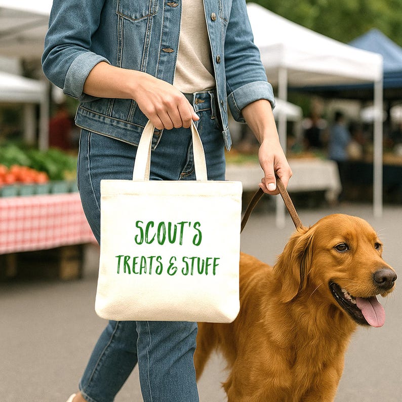 Cream-colored canvas tote bag with the custom text in green. A golden retriever dog is on a leash next to the person holding the bag.