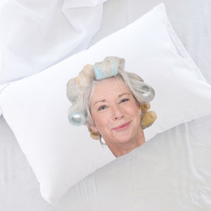 May include: A white pillow with a woman's face printed on it. The woman has grey hair in rollers and is smiling.