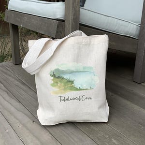 May include: A natural canvas tote bag featuring a watercolor landscape design with the text "Tidalwood Cove." The image depicts a serene scene with green, blue, and yellow hues. The bag has a sturdy handle for carrying.