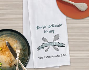 May include: White kitchen towel with the text "You're welcome in my kitchen when it's time to do the dishes" and a graphic of crossed silverware with a banner that says "Kitchen".