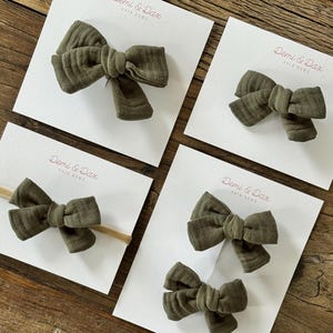 May include: Four olive green fabric hair bows attached to white display cards labeled Demi & Dax Hair Bows. The collection includes one large bow, one small bow, one bow on a tan elastic headband, and a pair of small bows on a single card.