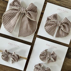 May include: Four hair bows with a brown and white gingham check pattern, each featuring a cream-colored piped edge. The bows are displayed on white cards labeled Demi & Dax Hair Bows, with one bow attached to a tan elastic headband.