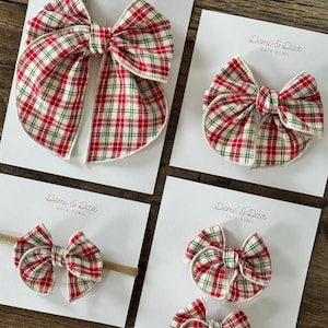 May include: Four hair bows featuring a red, white, and green plaid pattern on white display cards labeled Demi & Dax Hair Bows. The collection includes one large bow, one medium bow, one small bow on a tan headband, and two small bows.