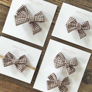 May include: Four brown and white gingham hair bows displayed on white cards labeled Demi & Dax Hair Bows. One bow is on a tan elastic headband, one is a single clip, and two are paired on a single card. The fabric features a small checkered pattern.