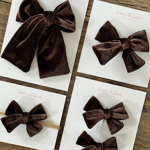 May include: Four dark brown velvet hair bows displayed on white branded cards labeled Demi & Dax Hair Bows. The collection includes one large bow, one medium bow, one small bow on a tan elastic headband, and a pair of small bows on a single card.