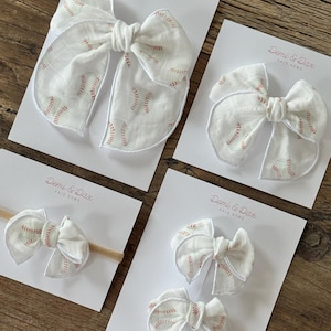 May include: White fabric hair bows with red baseball stitching detail, displayed on white cards. The bows come in various sizes, including a headband and clip-on styles. The Demi & Dax logo is printed on the cards.