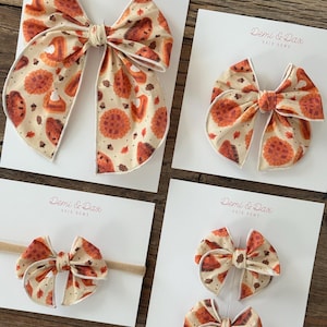 May include: Four hair bows in varying sizes featuring a cream-colored fabric with a repeating pattern of orange pumpkin pies, acorns, and autumn leaves. Each bow is attached to a white display card labeled with the text Demi & Dax Hair Bows.