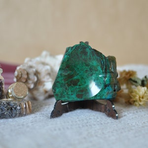 Può includere: Una lastra lucida di minerale crisocolla verde scuro e marrone su un supporto in legno scuro, accompagnata da due piccole fiale di vetro riempite di lavanda essiccata e petali di fiori, e una conchiglia bianca.