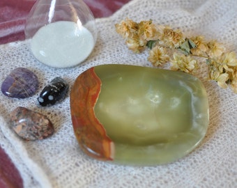 Puede incluir: Un plato de piedra pulida en verde y marrón con una forma suave y redondeada. Junto a él hay tres pequeñas piedras pulidas en morado, negro con puntos blancos y marrón moteado, además de un reloj de arena de cristal con arena blanca.