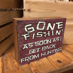 Op de afbeelding: Een houten bord met rode verf en witte letters met de tekst "Gone Fishin' As Soon As I Get Back From Hunting".