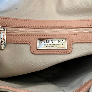 May include: Close-up of a light pink leather handbag interior. The zipper is closed, and the pull has a decorative "V" emblem. A silver rectangular tag reads "VALENTINA GENUINE LEATHER MADE IN ITALY".