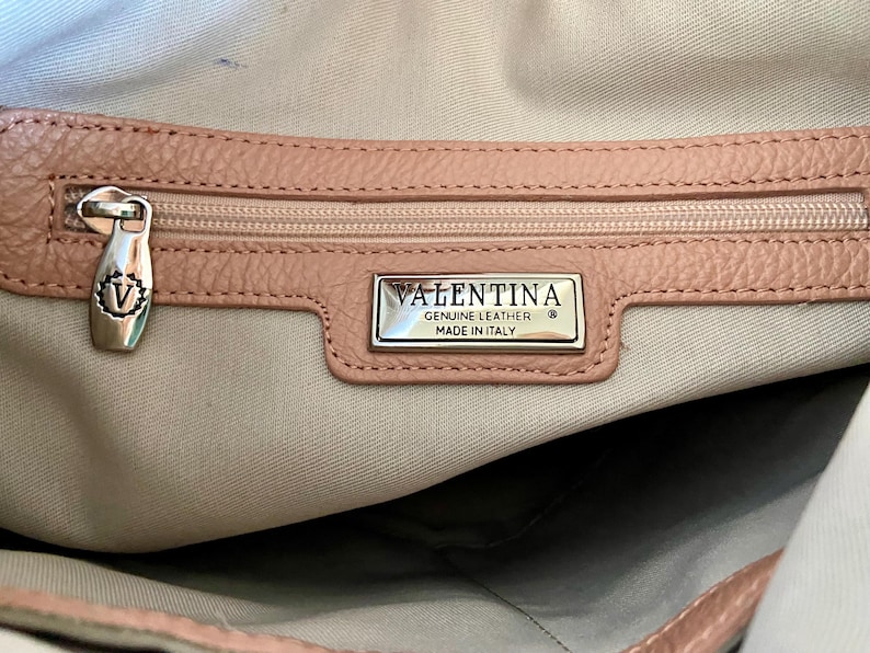 May include: Close-up of a light pink leather handbag interior. The zipper is closed, and the pull has a decorative "V" emblem. A silver rectangular tag reads "VALENTINA GENUINE LEATHER MADE IN ITALY".