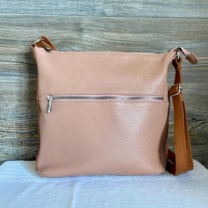May include: A blush pink leather crossbody bag with a brown adjustable strap. The bag has a front zipper pocket with a silver zipper and a matching silver zipper on the main compartment. The bag is sitting on a white surface.
