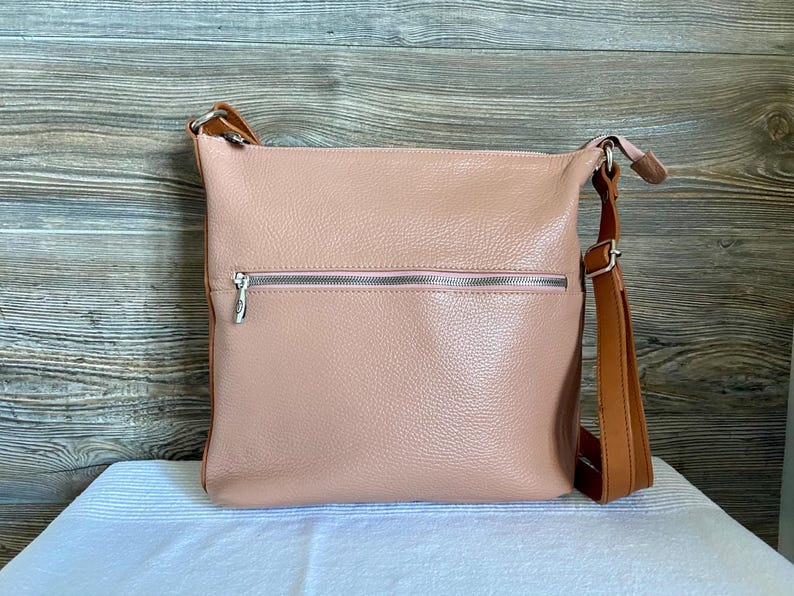 May include: A blush pink leather crossbody bag with a brown adjustable strap. The bag has a front zipper pocket with a silver zipper and a matching silver zipper on the main compartment. The bag is sitting on a white surface.