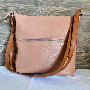 May include: A blush pink leather crossbody bag with a brown adjustable strap. The bag has a zippered pocket on the front and a matching pink zipper pull. The bag is sitting on a white surface.
