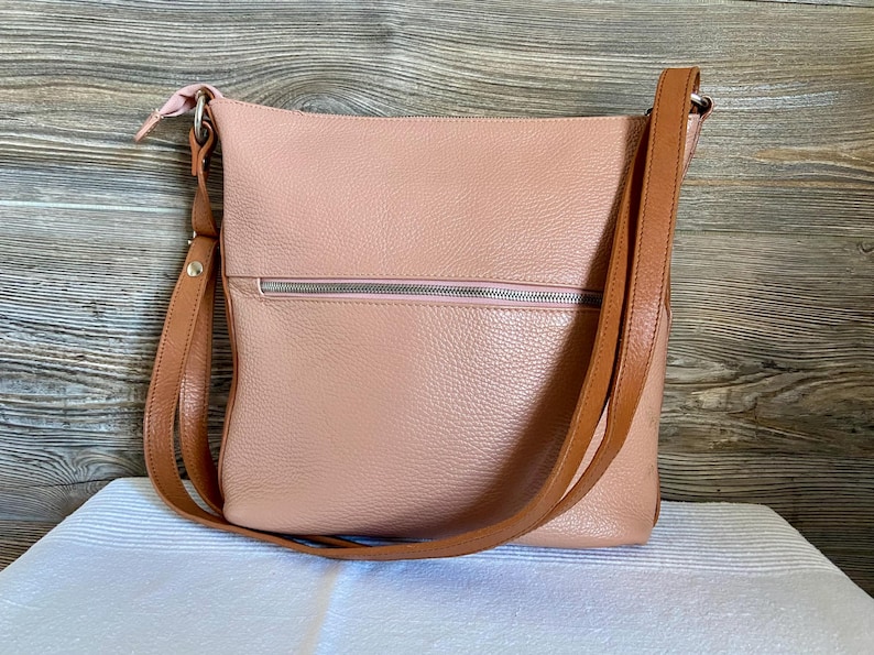 May include: A blush pink leather crossbody bag with a brown adjustable strap. The bag has a zippered pocket on the front and a matching pink zipper pull. The bag is sitting on a white surface.