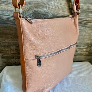 May include: A blush pink leather crossbody bag with a brown leather strap and trim. The bag has a zippered top and a front zippered pocket. The hardware is silver-toned. The bag is sitting on a white surface.