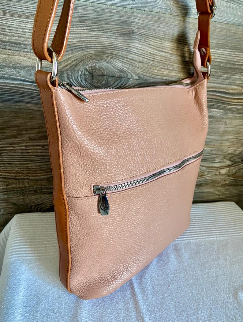 May include: A blush pink leather crossbody bag with a brown leather strap and trim. The bag has a zippered top and a front zippered pocket. The hardware is silver-toned. The bag is sitting on a white surface.