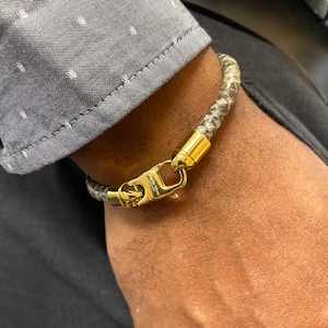 May include: A braided bracelet with a snakeskin pattern in shades of grey and tan. The bracelet has gold-coloured metal clasps and end caps. The clasp is engraved with the text "martin bine".