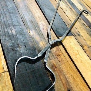 May include: A pair of black metal fireplace tongs with a twisted design. The tongs are lying on a wooden surface.