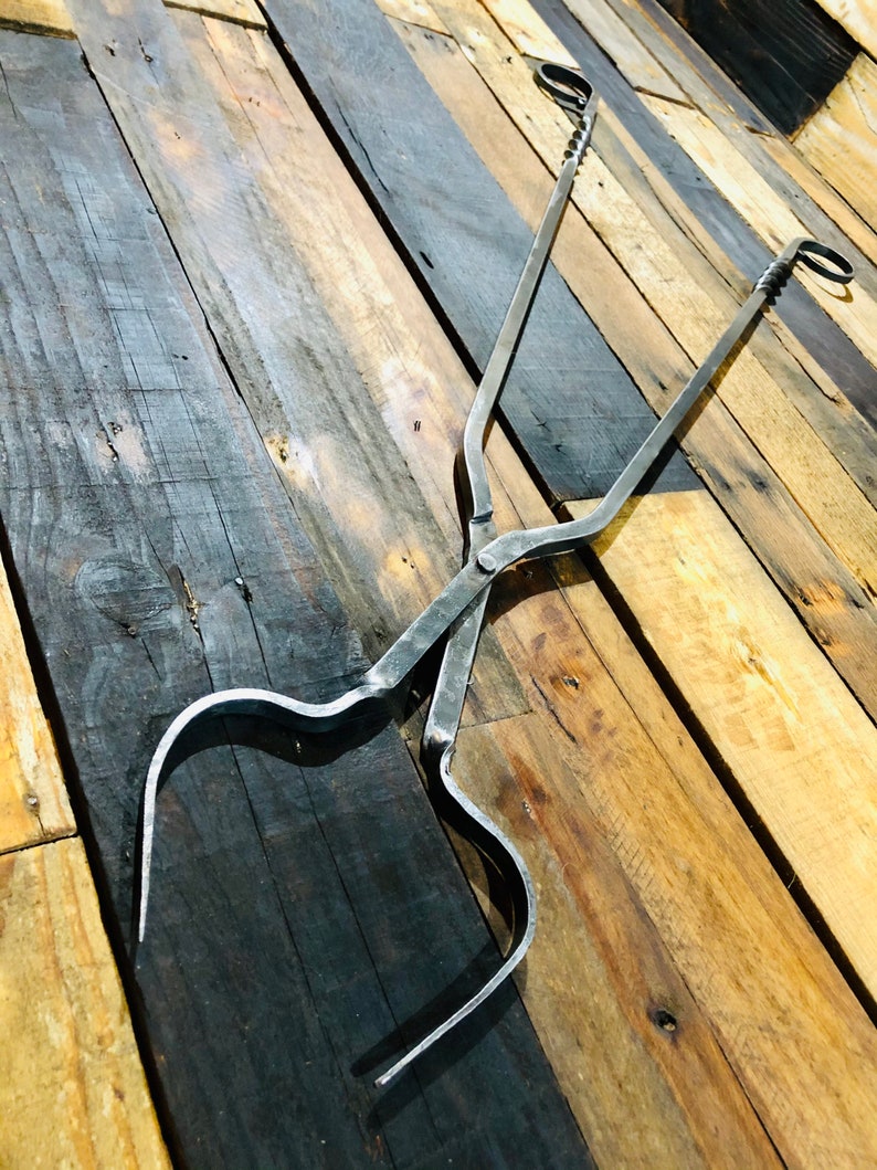 May include: A pair of black metal fireplace tongs with a twisted design. The tongs are lying on a wooden surface.