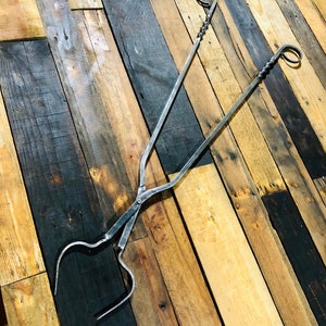 May include: A pair of black metal fireplace tongs with twisted handles and rounded ends. The tongs are lying on a wooden surface.