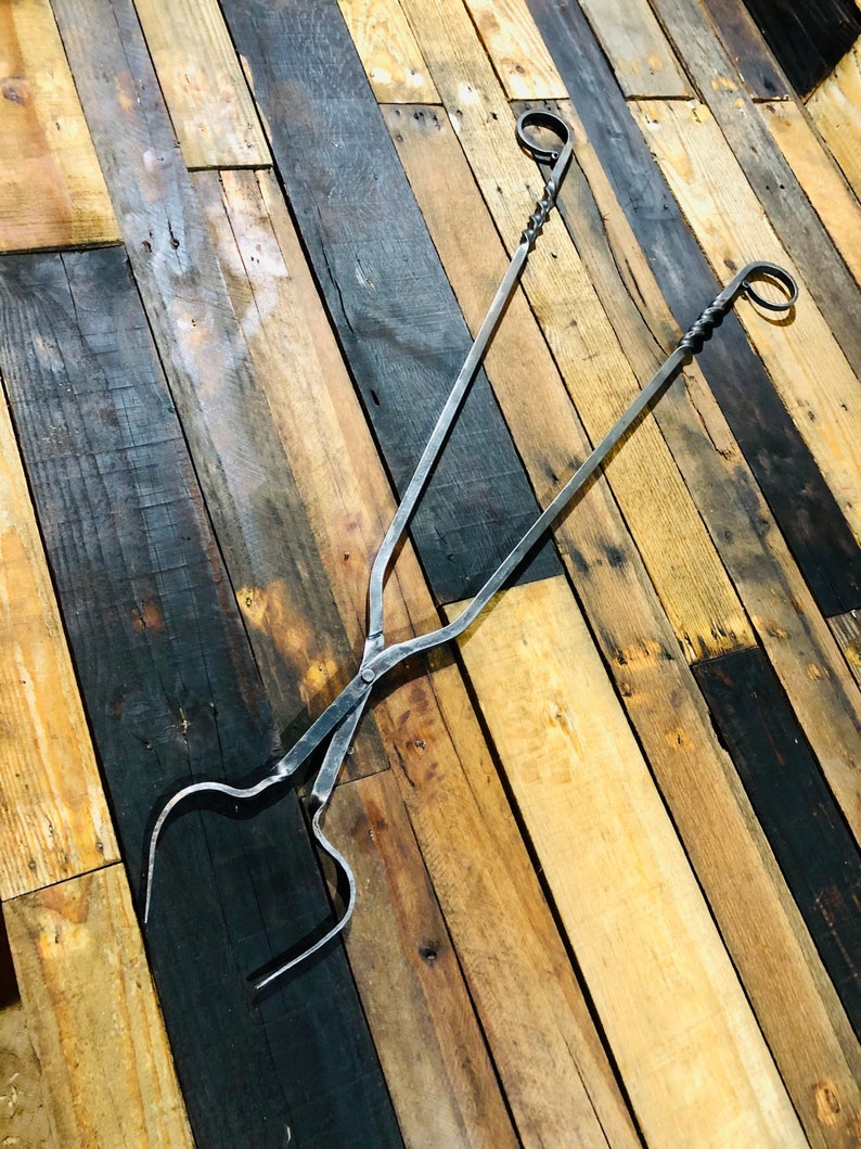 May include: A pair of black metal fireplace tongs with twisted handles and rounded ends. The tongs are lying on a wooden surface.