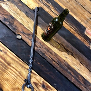 May include: A black metal bottle opener with a twisted handle and a ring at the end. The opener is lying on a wooden surface next to a green glass bottle with a label that says "Sonoma Cider".