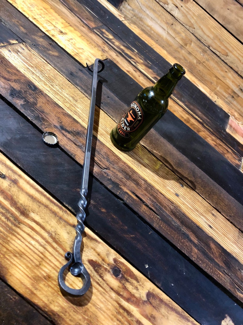 May include: A black metal bottle opener with a twisted handle and a ring at the end. The opener is lying on a wooden surface next to a green glass bottle with a label that says "Sonoma Cider".