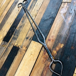 May include: A pair of black metal fireplace tongs with a twisted handle and heart-shaped ends. The tongs are lying on a wooden surface.