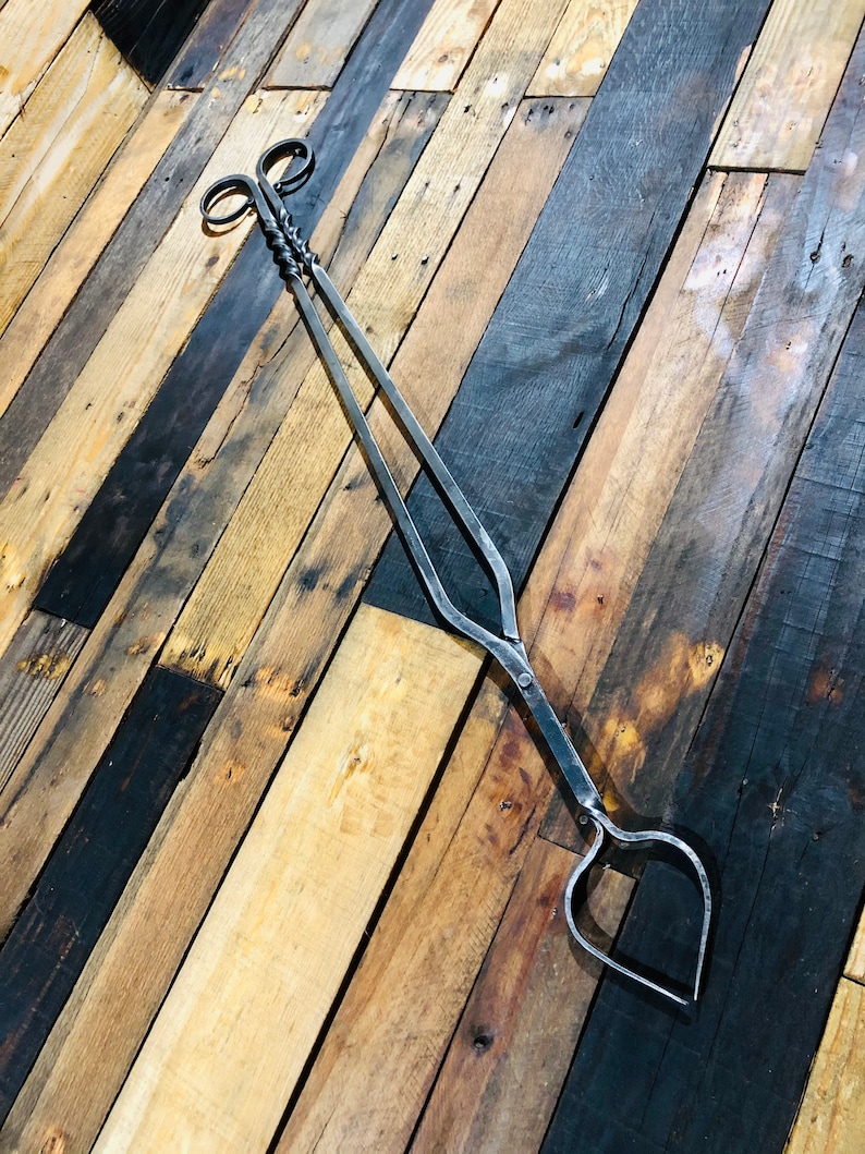 May include: A pair of black metal fireplace tongs with a twisted handle and heart-shaped ends. The tongs are lying on a wooden surface.