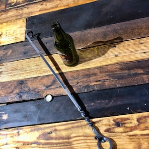 May include: A long, twisted metal skewer with a loop at each end. The skewer is lying on a wooden surface with a green glass bottle.