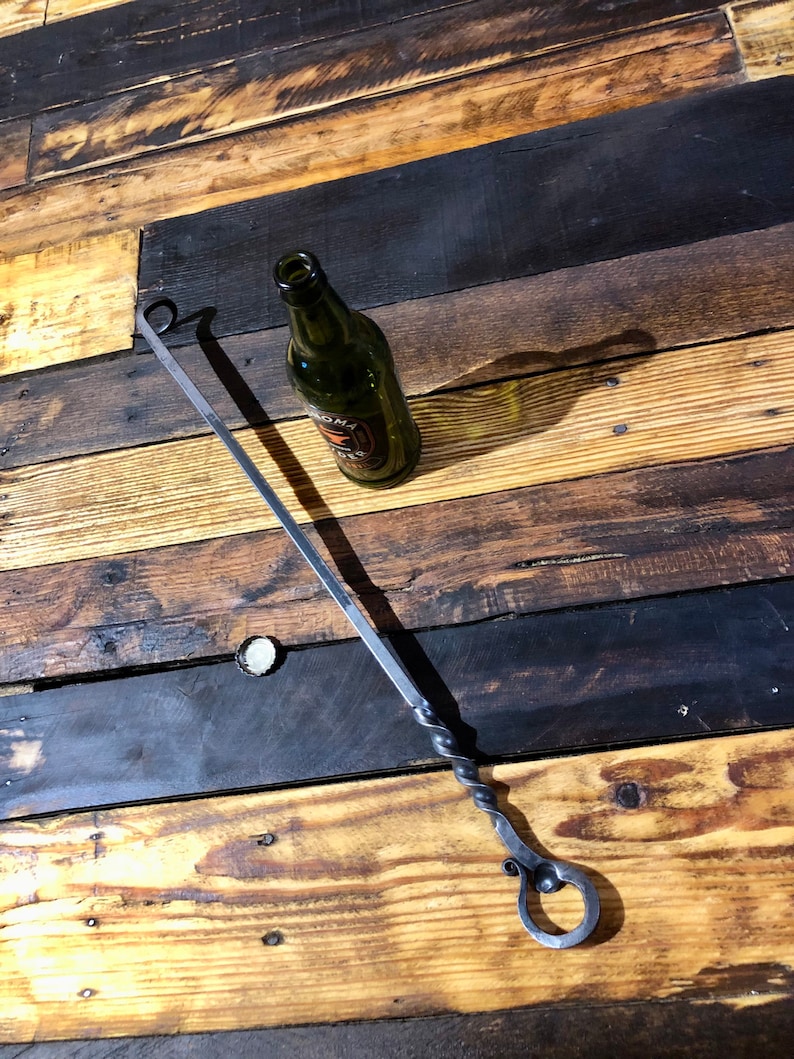May include: A long, twisted metal skewer with a loop at each end. The skewer is lying on a wooden surface with a green glass bottle.