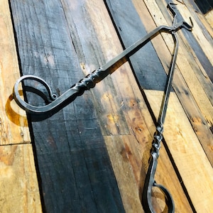 May include: A black metal fireplace tool with a twisted handle and two hooks. The tool is hanging on a wooden wall.
