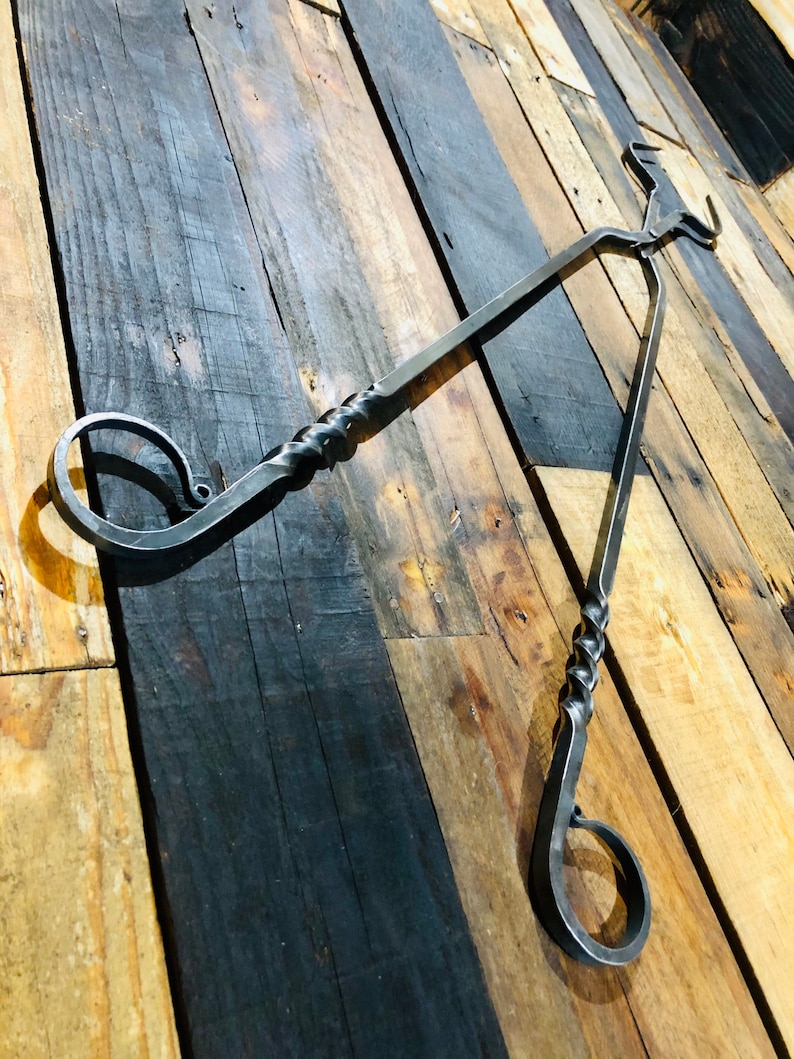 May include: A black metal fireplace tool with a twisted handle and two hooks. The tool is hanging on a wooden wall.