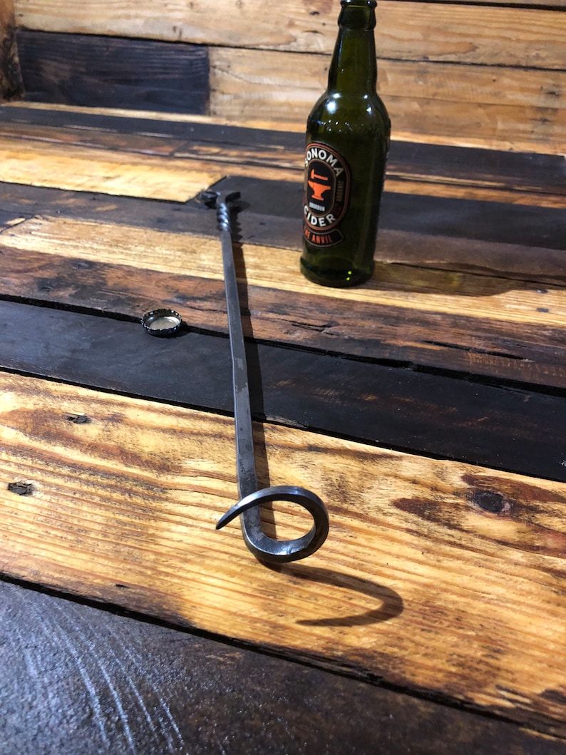 May include: A black metal fireplace poker with a curved handle. The poker is lying on a wooden surface. A bottle of Sonoma Cider is in the background.