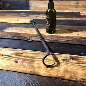 May include: A black metal fireplace poker with a hook on the end. The poker is lying on a wooden surface next to a green glass bottle with a label that says "Sonoma Cider".