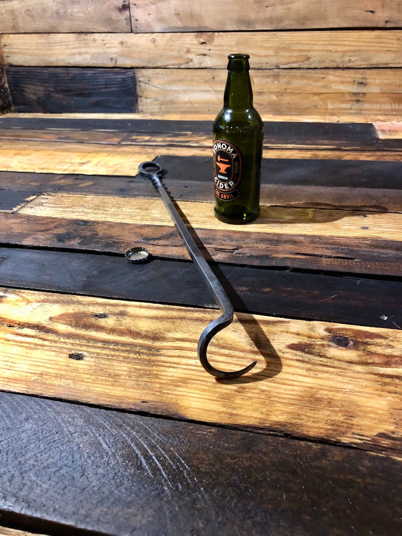 May include: A black metal fireplace poker with a hook on the end. The poker is lying on a wooden surface next to a green glass bottle with a label that says "Sonoma Cider".