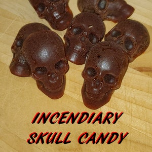May include: A close-up of several chocolate skull-shaped sweets on a wooden surface. The sweets are brown and have a glossy finish. The text "Blazing Dragon Incendiary Skull Candy" is printed on the surface.