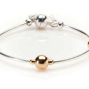 Puede incluir: Pulsera de plata con detalles dorados. La pulsera presenta una banda plateada con tres esferas doradas y un cierre plateado con cierre magnético. La pulsera está diseñada con un estilo minimalista.