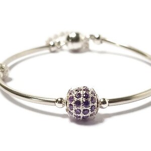 Pode incluir: Uma pulseira prateada com um berloque esférico central coberto por pequenas pedras preciosas roxas. A pulseira tem um design simples e elegante com um fecho e está sobre um fundo branco.