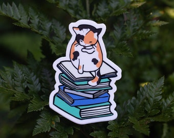 May include: A sticker featuring a calico cat sitting atop a stack of books. The cat has orange, black, and white markings. The books are blue and teal with white pages. The sticker is set against a backdrop of green foliage.