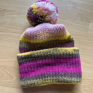 May include: A striped knitted beanie with a multi-coloured pom pom. The beanie is pink, yellow and brown.